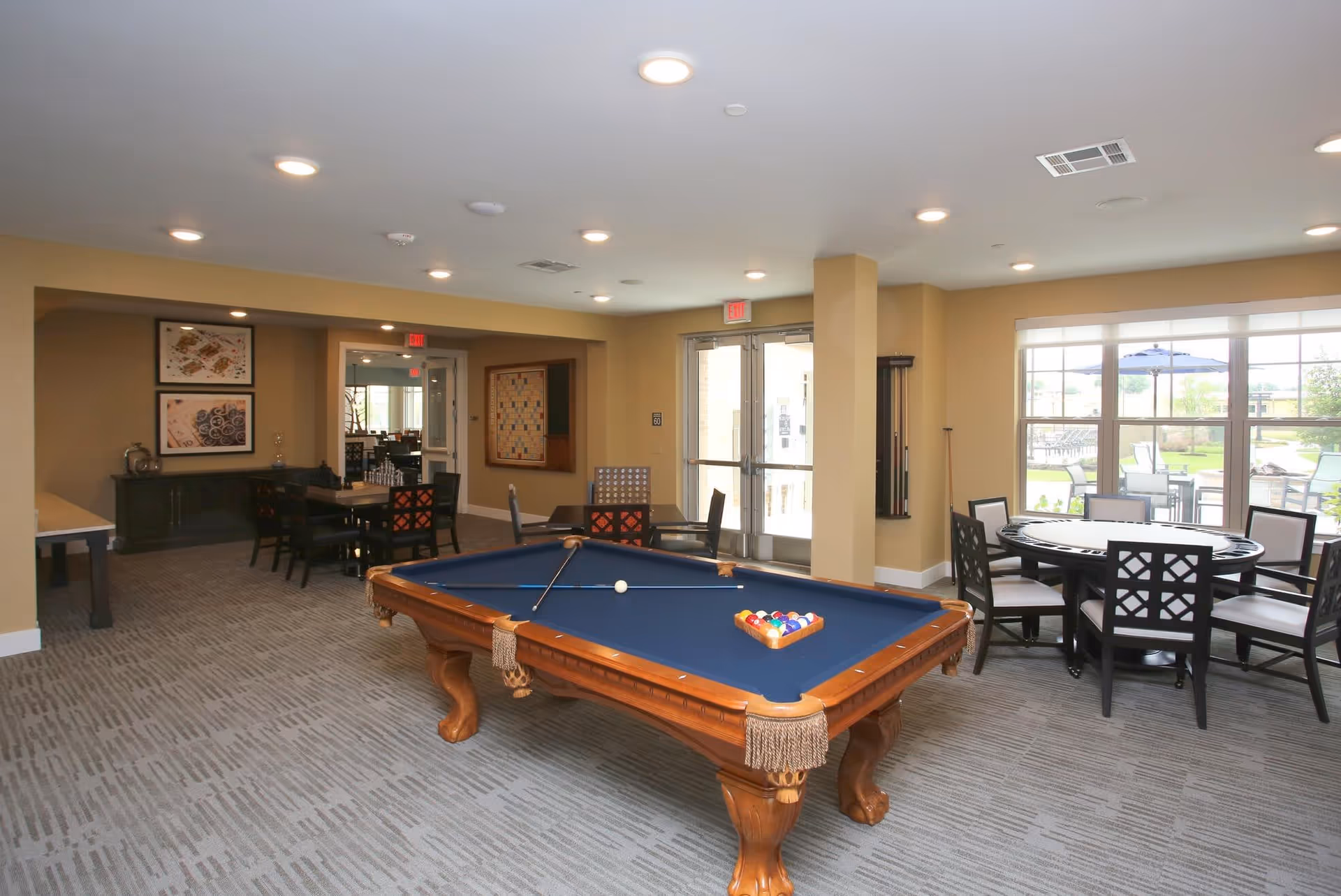 Bright communal game room with a pool table in the center, seating and a round card table by large windows.