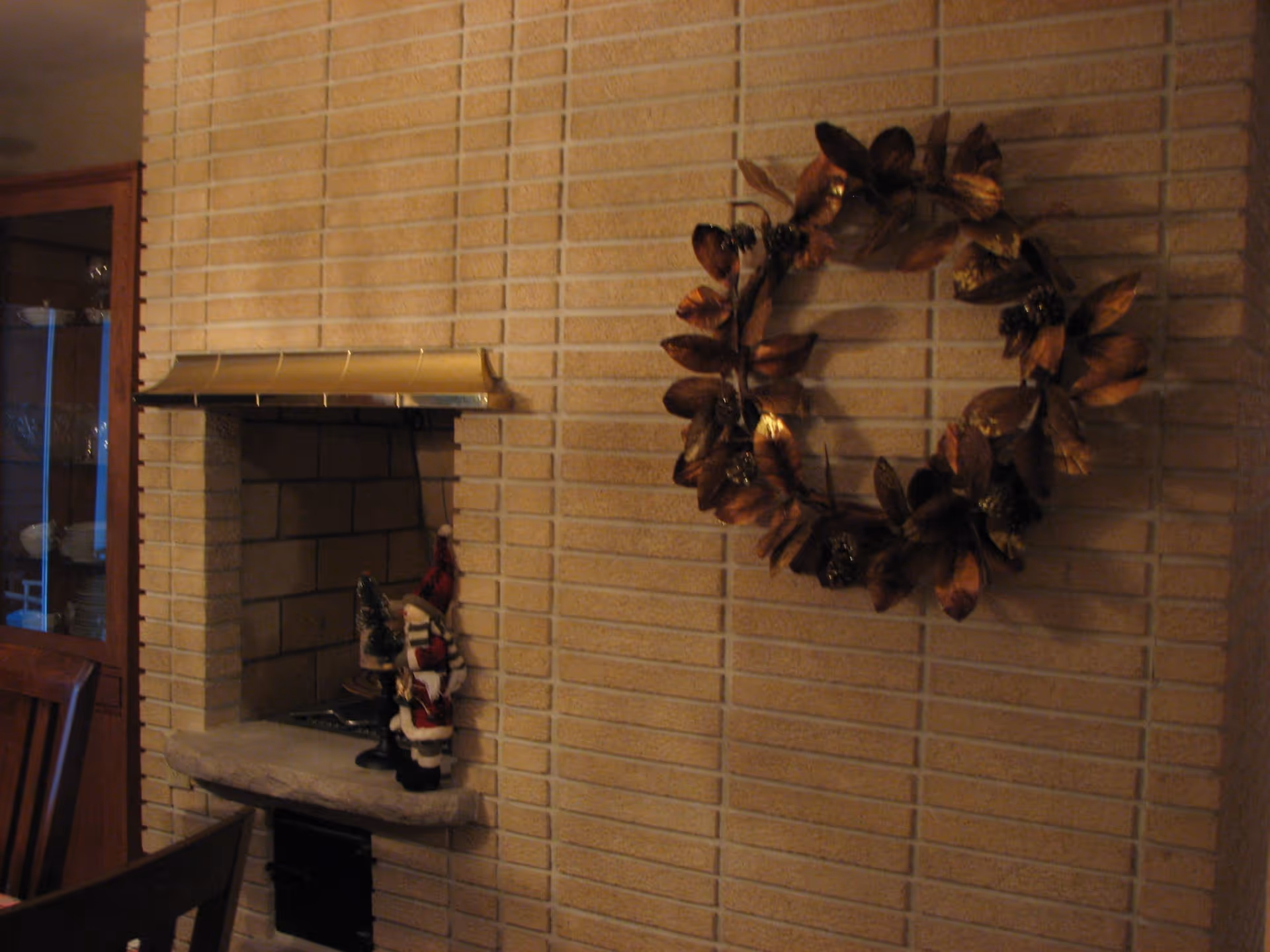 A cozy corner of a room with a brick fireplace featuring a brass hood. On the fireplace hearth, there are small decorative figurines including a Santa Claus. To the right on the brick wall, there is a decorative wreath made of brown leaves and pinecones. Part of a wooden dining chair and a glass-fronted cabinet with dishes are visible on the left side.