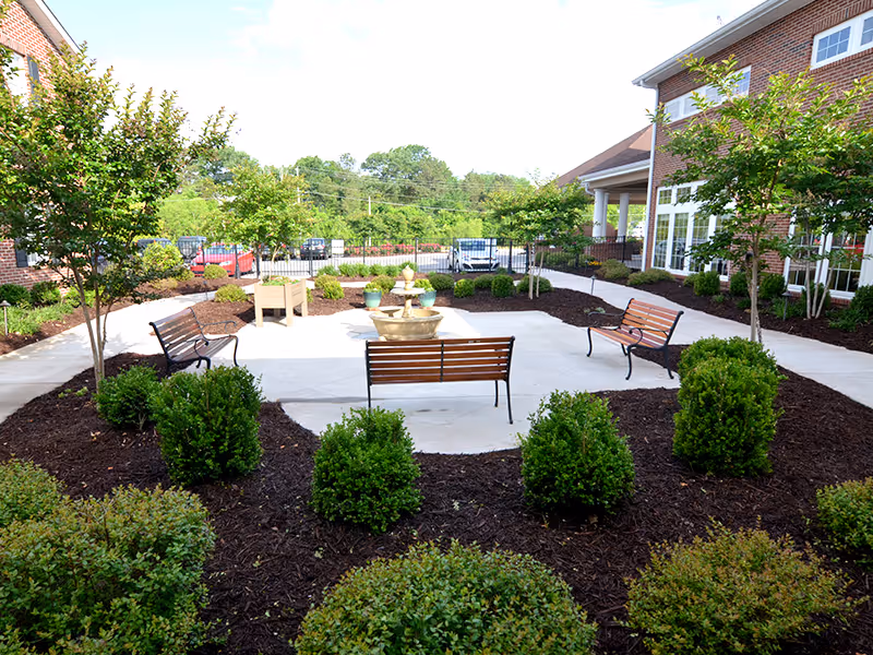 Courtyard with benches and a small fountain surrounded by landscaped shrubs and brick buildings.