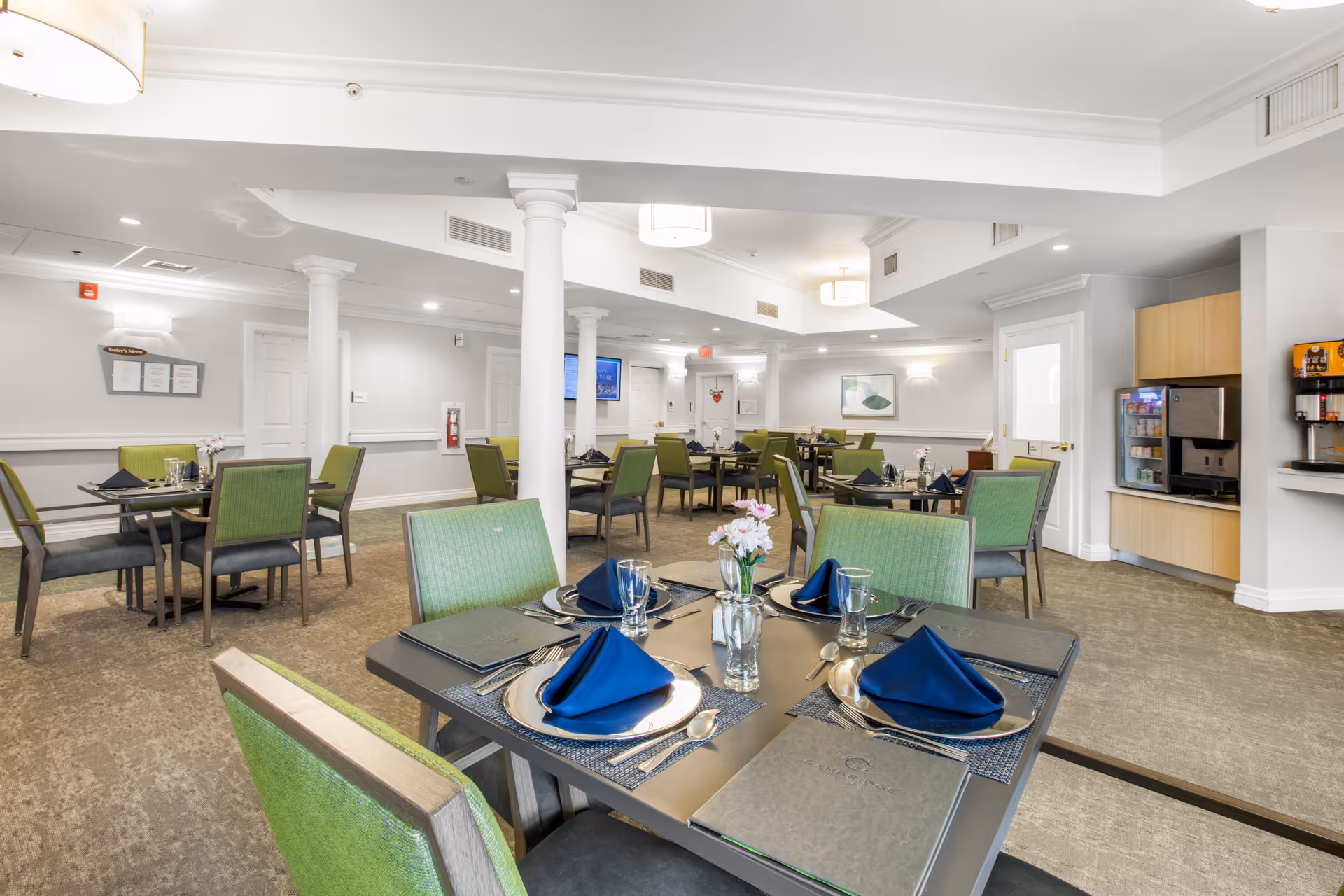 A bright and spacious dining room with multiple tables set for meals. Each table has green cushioned chairs, silver plates, blue folded napkins, glasses, and silverware. The room features white walls, columns, ceiling lights, and a beverage station with a coffee machine and a refrigerated drink dispenser in the corner.