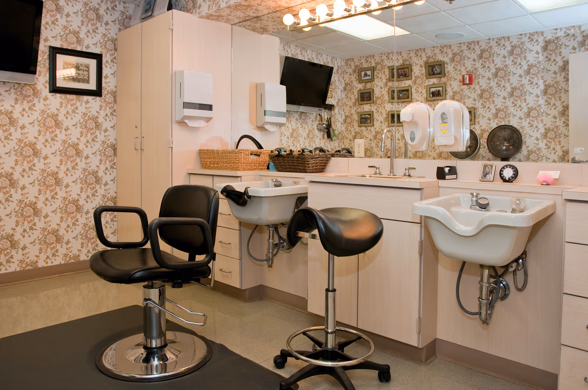 Interior of a salon or grooming area with two black salon chairs, two white sinks, light wood cabinetry, floral wallpaper, a large mirror, and wall-mounted soap and paper towel dispensers.