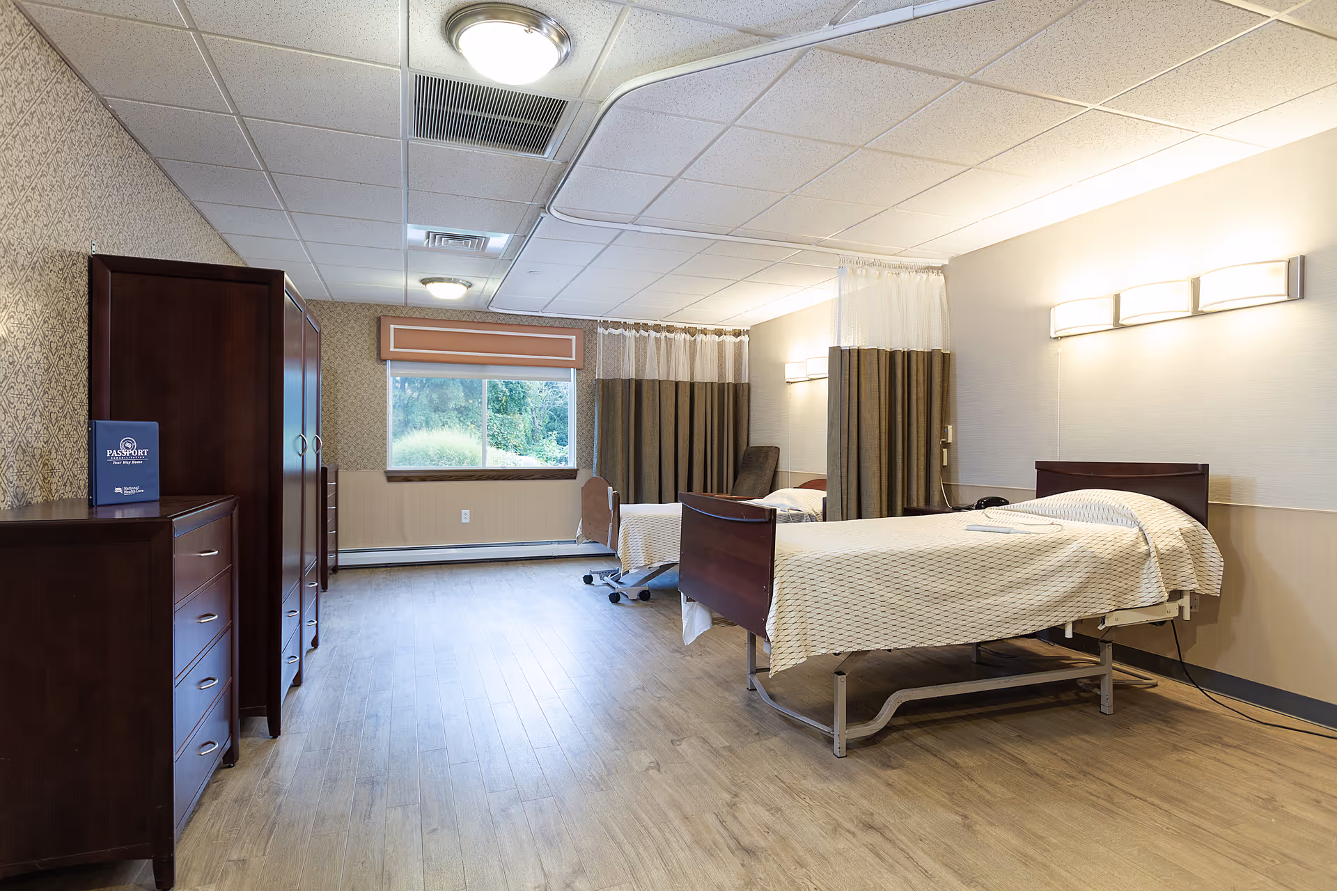 A spacious room in a senior living facility with two hospital-style beds separated by privacy curtains. The room has wooden furniture including dressers and wardrobes, a large window with a view of greenery outside, and soft lighting fixtures on the walls and ceiling. The floor is covered with wood-like vinyl flooring.