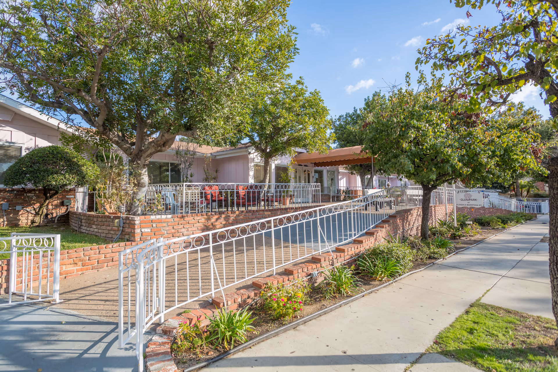 Front exterior of a senior living facility with a wheelchair ramp, brick planter walls, trees and a covered entrance.