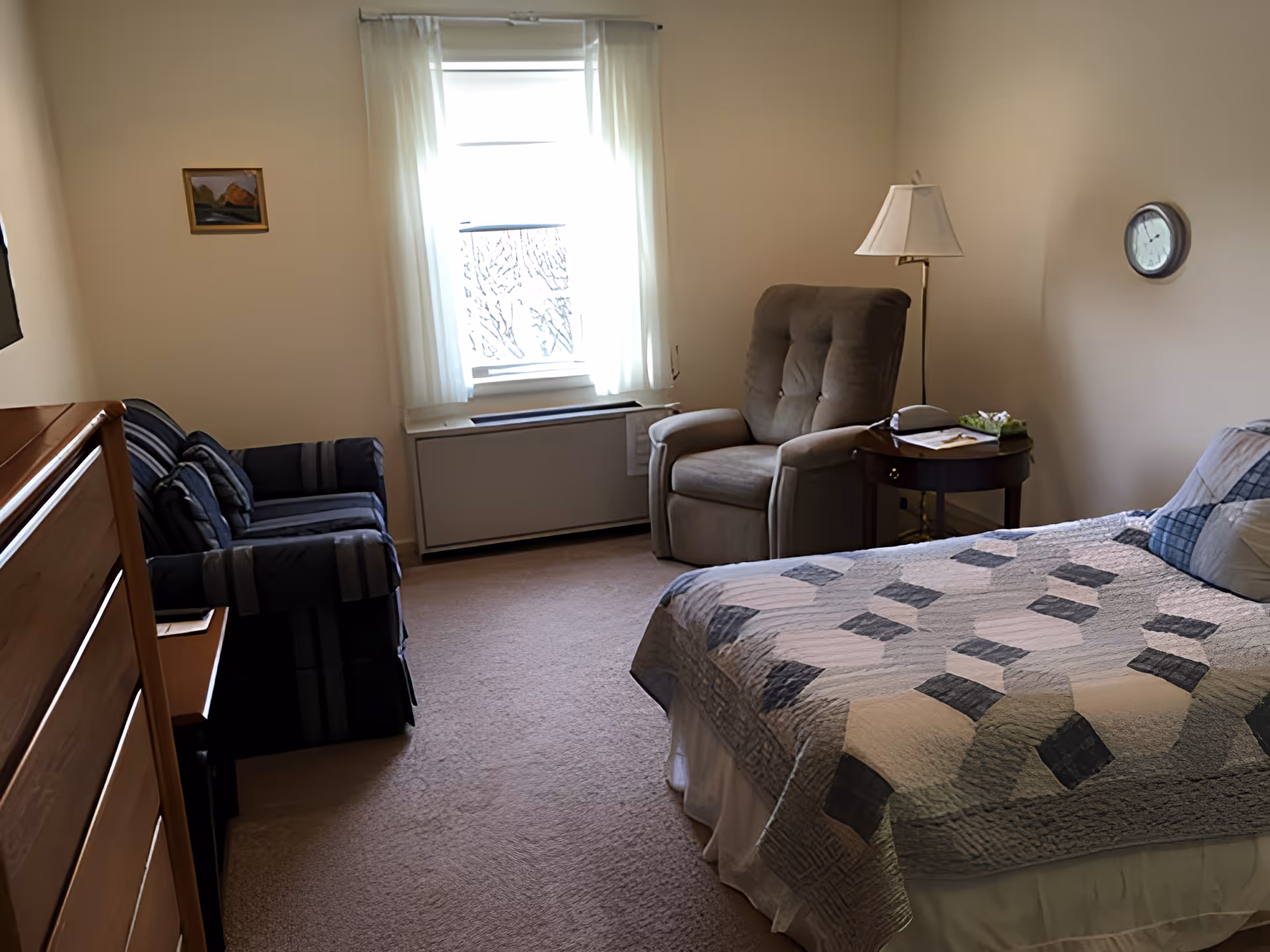 A cozy bedroom with a bed covered in a patterned quilt, a cushioned armchair next to a small wooden side table with a lamp, a striped loveseat, a wooden dresser, a window with sheer white curtains, a small framed painting on the wall, and a round wall clock.