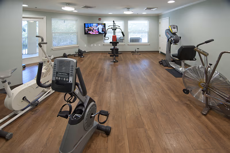 A senior living facility exercise room with various fitness equipment including stationary bikes, a multi-function weight machine, and an exercise fan. The room has wood flooring, light-colored walls, three windows with blinds, a wall-mounted TV, and ceiling lights.
