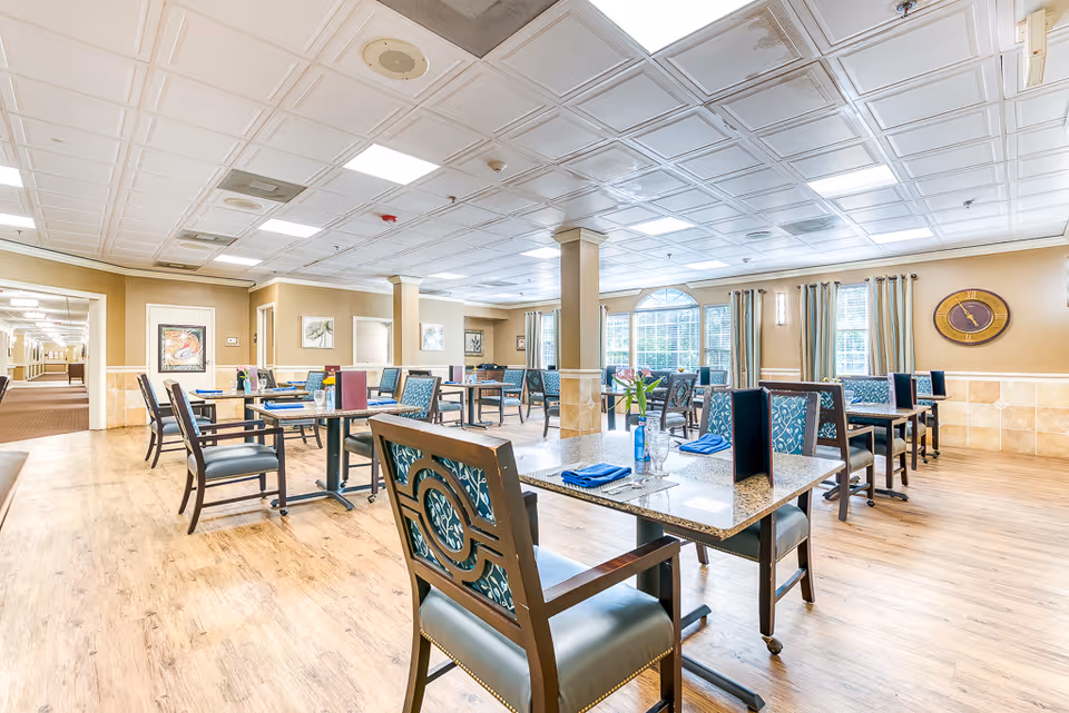 A bright and spacious dining room in a senior living facility with multiple tables and chairs arranged neatly. Each table has place settings with blue napkins, menus, and a small vase with flowers. The room features large windows with curtains, light-colored walls, and a wooden floor. There is a large clock on the wall and framed artwork decorating the space.