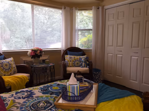 Cozy bedroom with a bed covered in colorful floral bedding, two armchairs by a large window, and a closed closet.