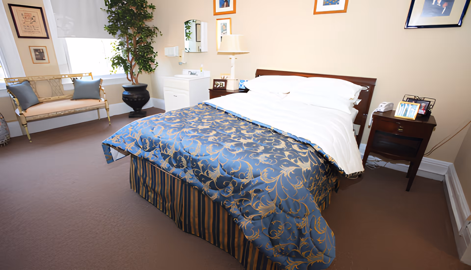 A bedroom in an assisted living facility featuring a neatly made bed with white sheets and a blue and gold patterned bedspread. There is a wooden nightstand with a lamp, framed photos, and a telephone. A small white sink with a mirror is mounted on the wall. A cushioned bench with two blue pillows is placed near a window with white blinds. The walls are decorated with framed pictures, and a large potted plant is in the corner.