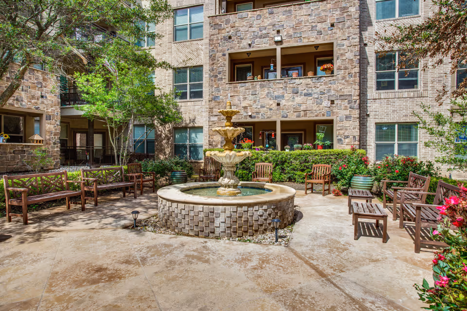 Outdoor courtyard area with a central three-tier stone fountain surrounded by wooden benches and chairs. The courtyard is bordered by a stone and brick building with windows and balconies, and there are green shrubs, potted plants, and trees providing a natural setting.