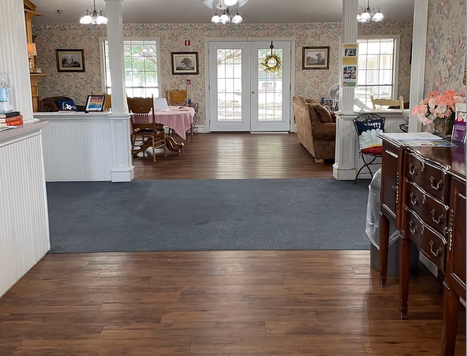 Interior view of a senior living facility common area with wooden flooring and a carpeted section. The room features floral wallpaper, two white pillars, a wooden sideboard with flowers and papers on the right, a small table with a pink tablecloth and wooden chairs in the background, and a brown couch on the right side near large windows and glass doors with a wreath hanging on the door.