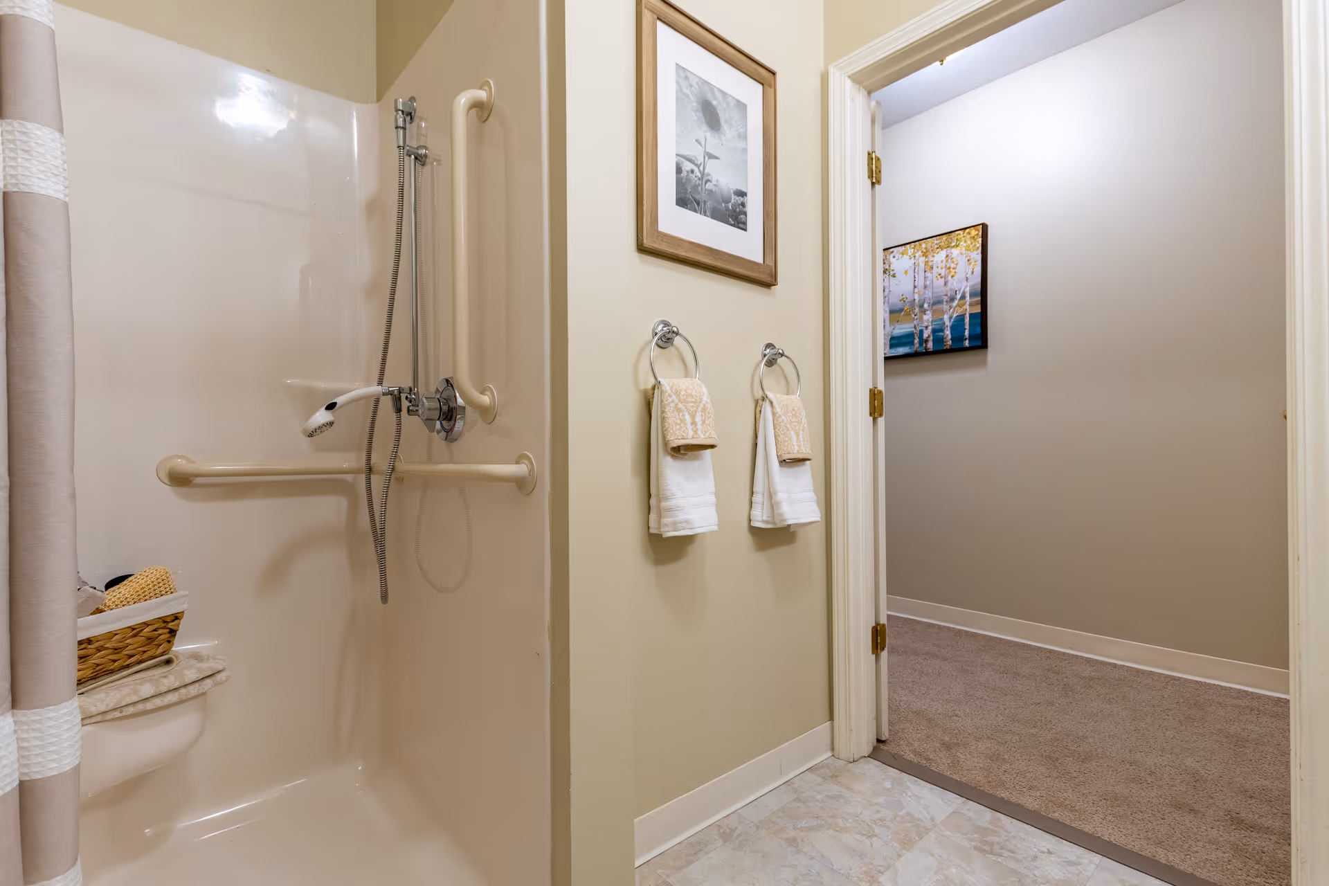 A bathroom with a beige shower area equipped with grab bars and a handheld showerhead. There is a basket with toiletries on a small shelf inside the shower. Two towel rings with beige and white towels hang on the wall outside the shower. The bathroom floor is tiled, and an open doorway leads to a carpeted hallway with a framed picture of birch trees on the wall.