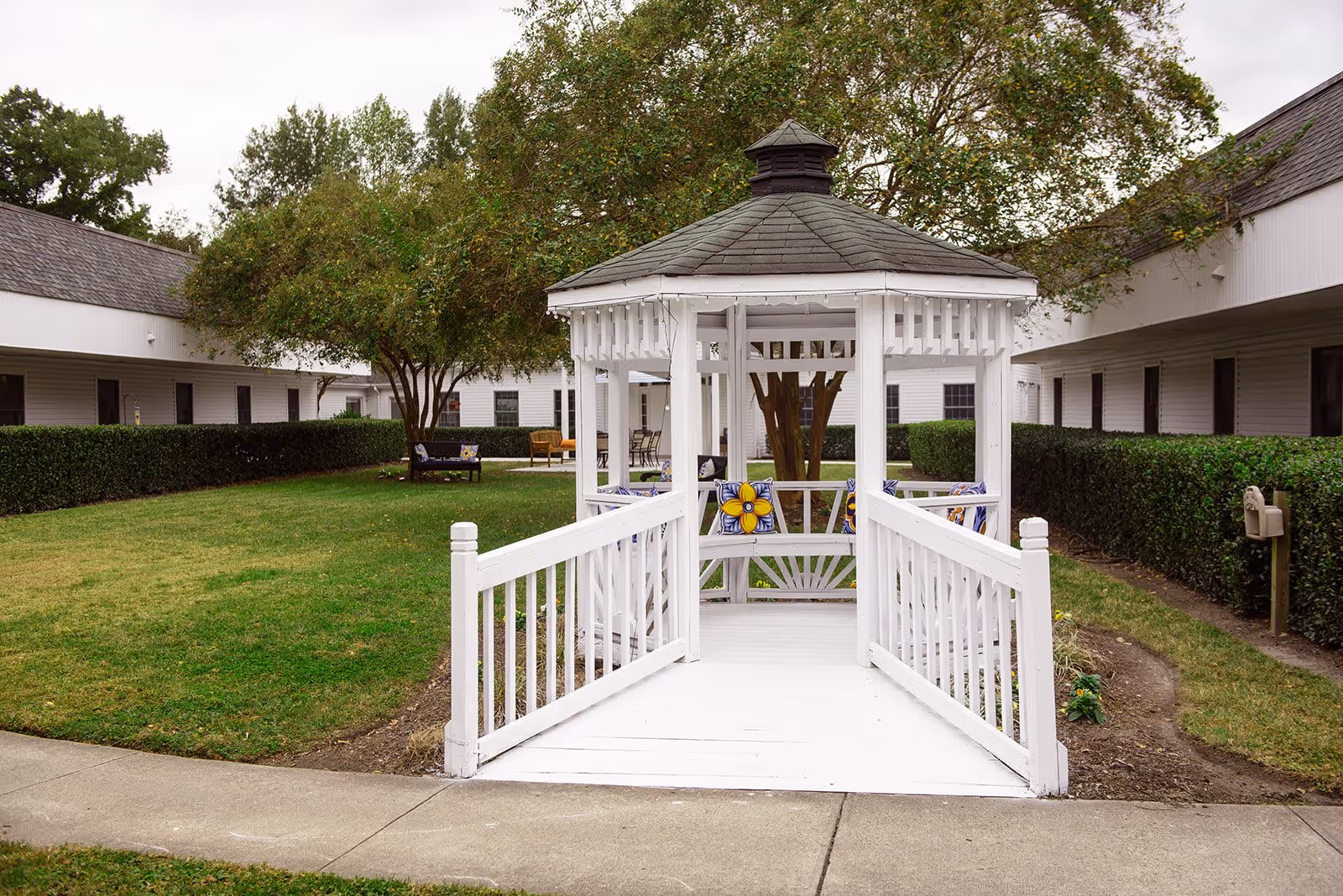 A white wooden gazebo with a shingled roof is situated in the center of a grassy courtyard surrounded by single-story buildings. The gazebo has a ramp leading up to it and is decorated with colorful flower-patterned cushions on the benches inside. Trees and trimmed hedges line the courtyard, and there are outdoor chairs and benches placed around the area.