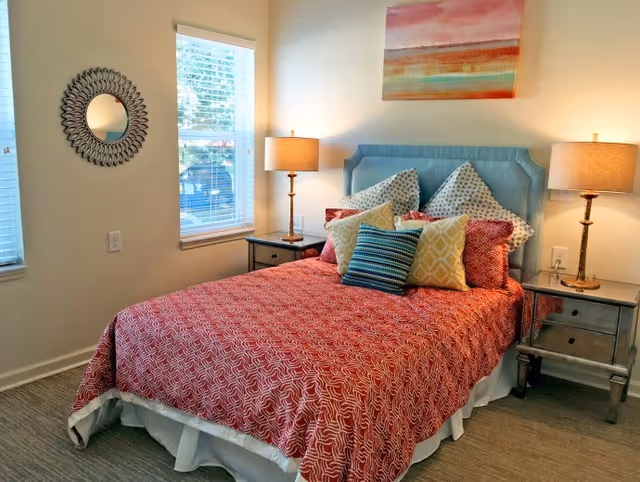 A bedroom with a bed covered in a red patterned bedspread and multiple decorative pillows in various colors and patterns. The bed has a light blue upholstered headboard. There are two bedside tables with matching lamps on each side of the bed. A round decorative mirror hangs on the wall to the left, and a colorful abstract painting is above the headboard. Two windows with white blinds allow natural light into the room.