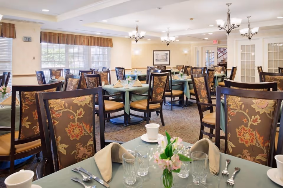 A well-lit dining room with multiple tables covered in green tablecloths, each set with beige napkins, cups, glasses, and silverware. The chairs have dark wooden frames with floral patterned upholstery. There are windows with striped valances letting in natural light, and chandeliers hanging from the ceiling. A small floral centerpiece is on the table in the foreground.