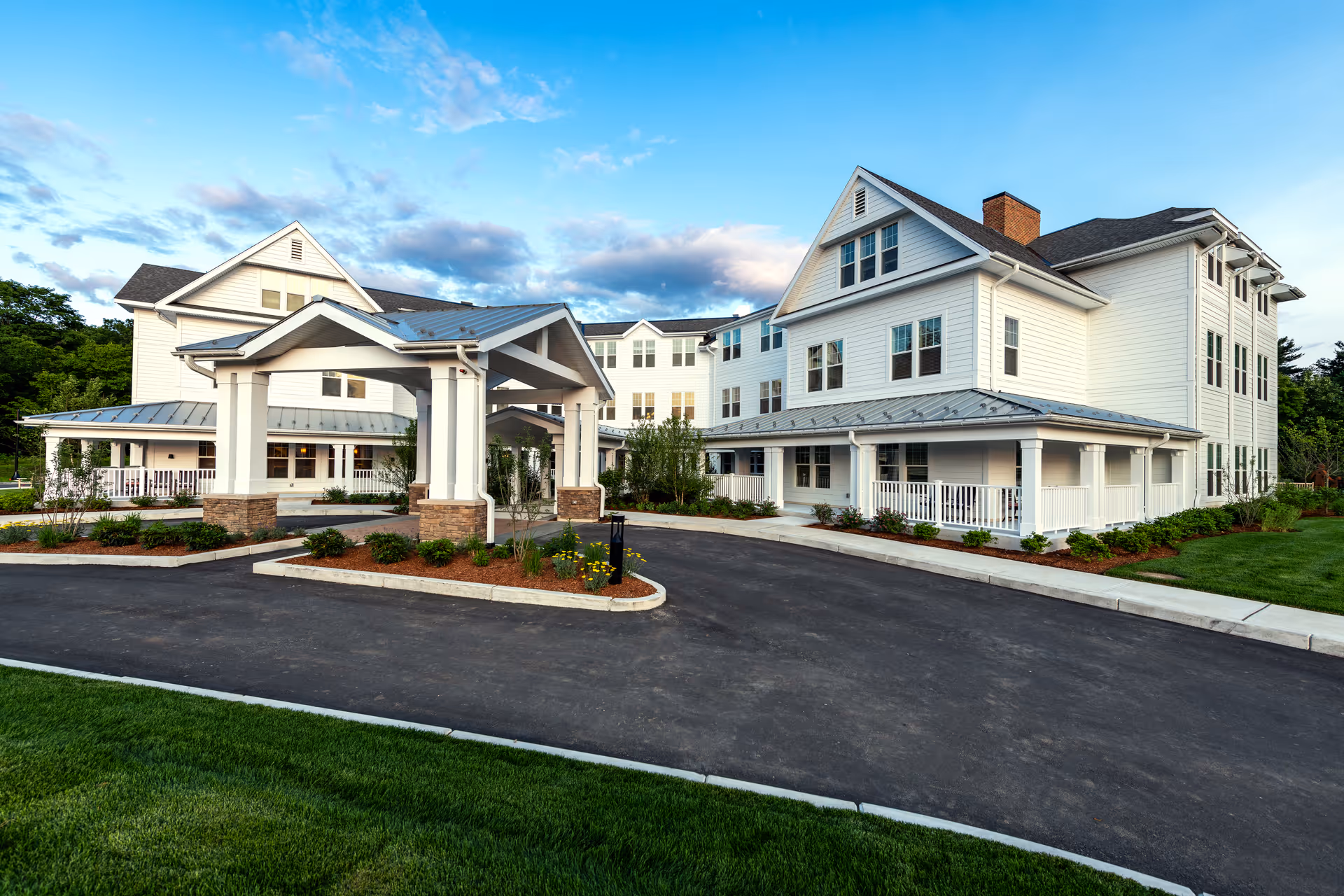 Bright white multi-story senior living building with a covered entrance/porte-cochere, wraparound porch, paved driveway and landscaped grounds under a blue sky.