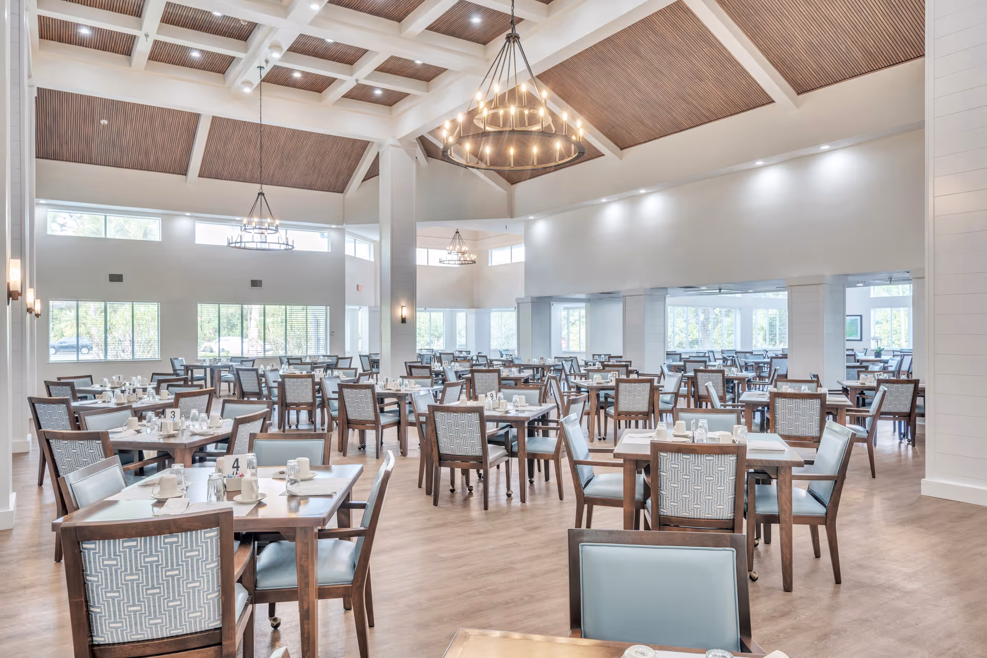 A spacious and well-lit dining room with multiple wooden tables and chairs arranged neatly. The room features large windows allowing natural light to fill the space, high ceilings with wooden beams, and modern chandeliers hanging from the ceiling. Each table is set with white cups, saucers, and napkins, creating an inviting atmosphere.