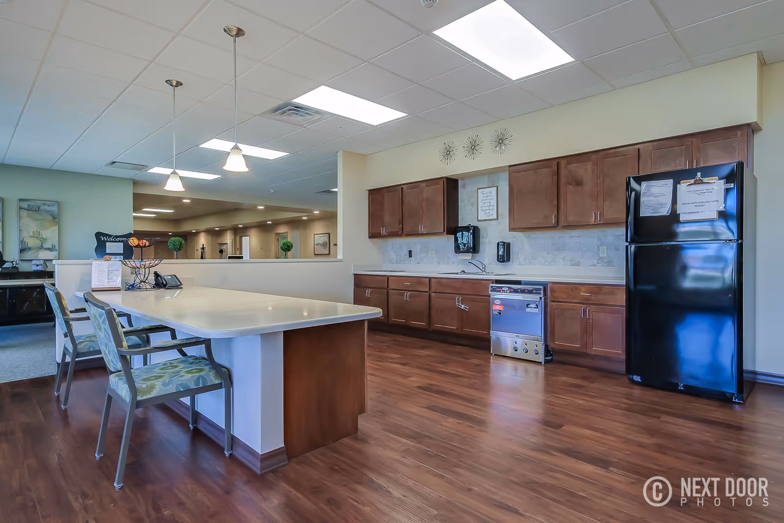 A spacious kitchen area in an assisted living facility featuring wooden cabinets, a black refrigerator with notices attached, a dishwasher, and a large island with two chairs. The room has wood flooring, pendant lights hanging from the ceiling, and a view into a hallway with decorative plants and artwork.