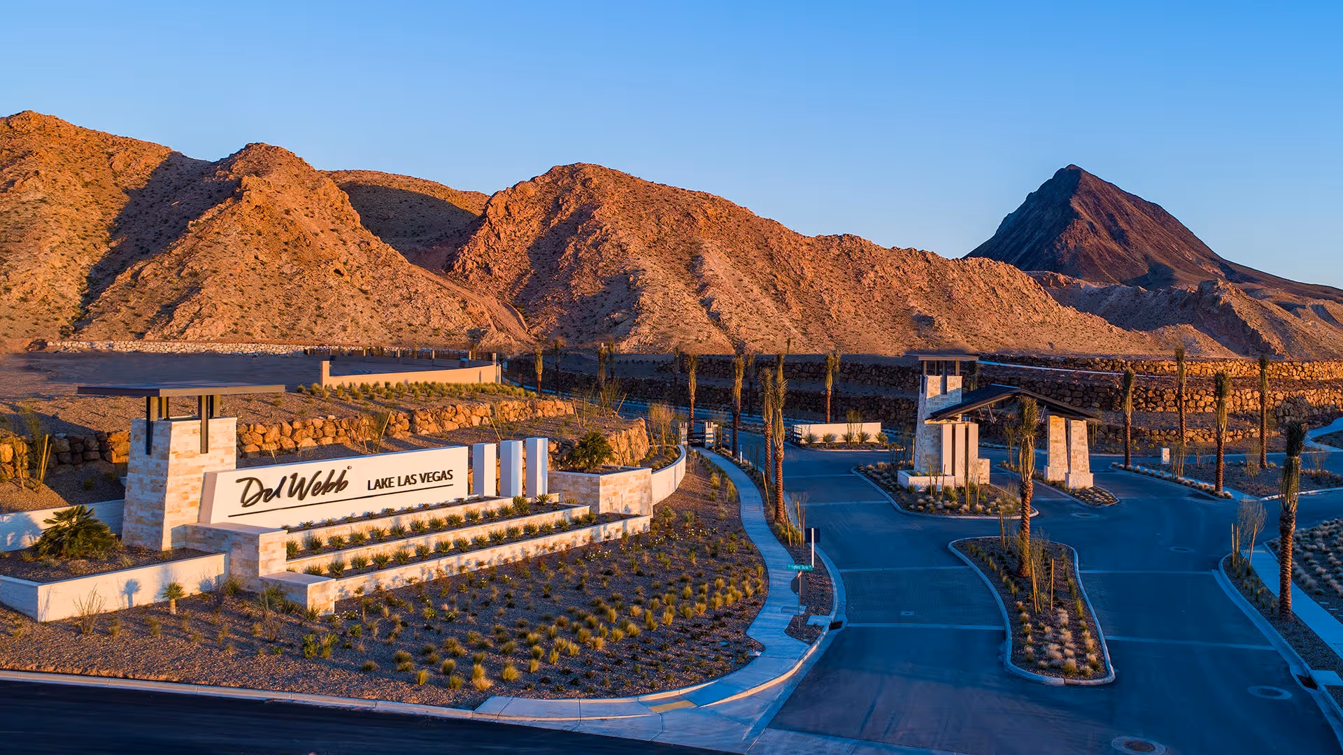 Entrance to Del Webb Lake Las Vegas retirement community with a large sign displaying the community name, surrounded by desert landscaping and mountains in the background under a clear blue sky.