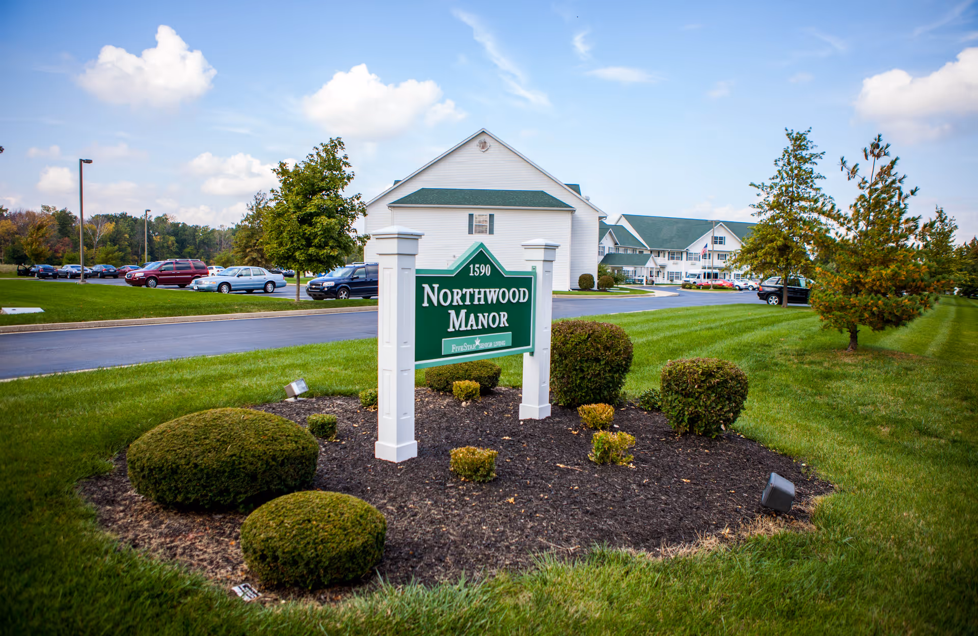 A green and white sign reading "Northwood Manor" sits in a landscaped lawn with the facility building and parked cars visible behind it.