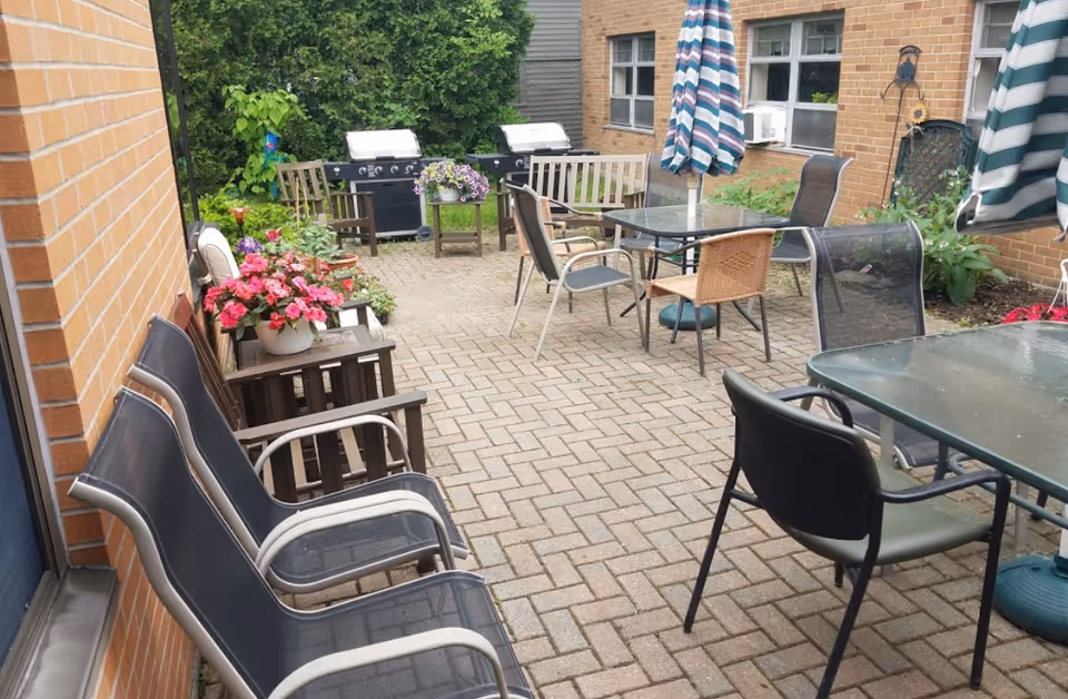 Outdoor patio area with multiple chairs and tables, two striped umbrellas, potted flowers on a small table, two grills in the background, and greenery along the edges near a brick building.