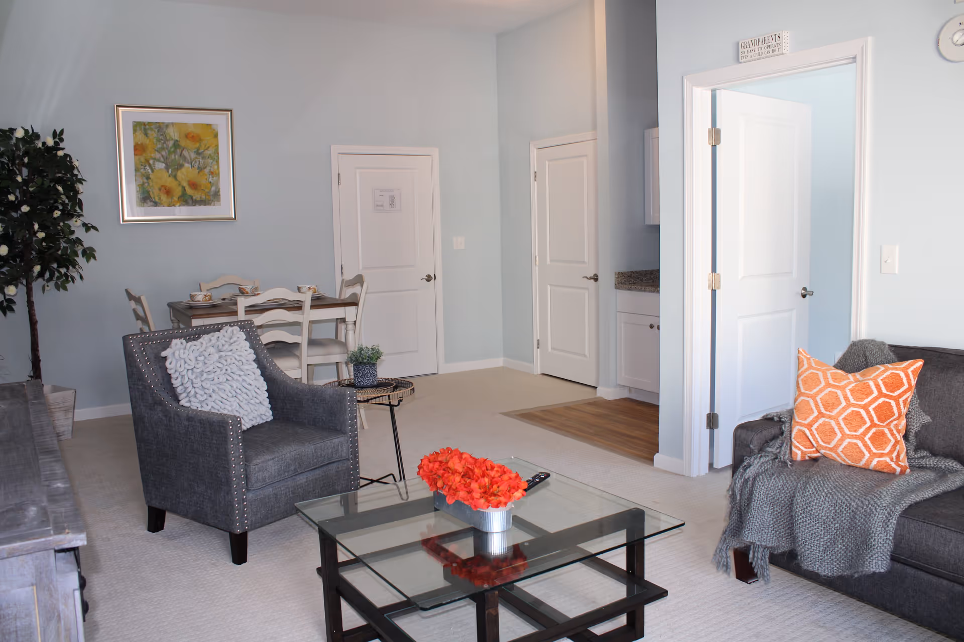 A cozy senior living room with a gray armchair featuring a textured white pillow, a gray sofa with an orange patterned pillow and a gray throw blanket, and a glass coffee table with a red flower centerpiece. In the background, there is a small dining table with four chairs, a framed floral painting on the wall, and two closed white doors. The walls are painted light blue, and there is a small plant on a side table next to the armchair.