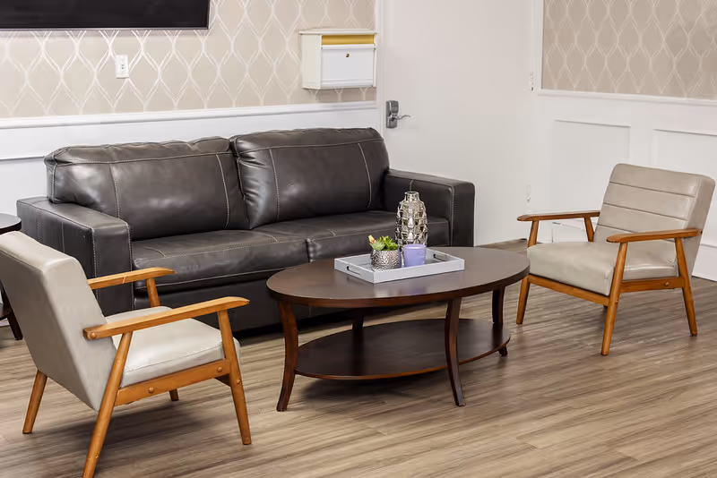 A cozy seating area with a dark brown leather sofa, two light beige armchairs with wooden armrests, and a dark wooden oval coffee table with decorative items on top. The room has light wood flooring and patterned wallpaper with a white door in the background.