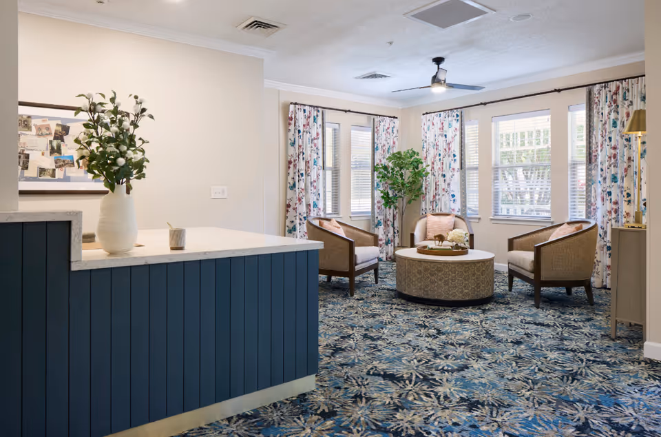 A cozy sitting area in a senior living facility with three cushioned armchairs arranged around a round coffee table. The room has large windows with floral curtains, a ceiling fan, and a blue patterned carpet. A blue reception desk with a white marble countertop and a vase with flowers is visible in the foreground.