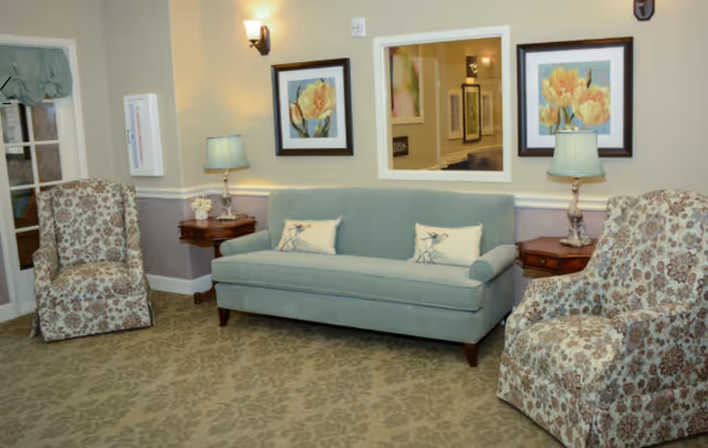 A seating area with a blue sofa flanked by two patterned armchairs, side tables, lamps, and framed floral artwork on the wall.