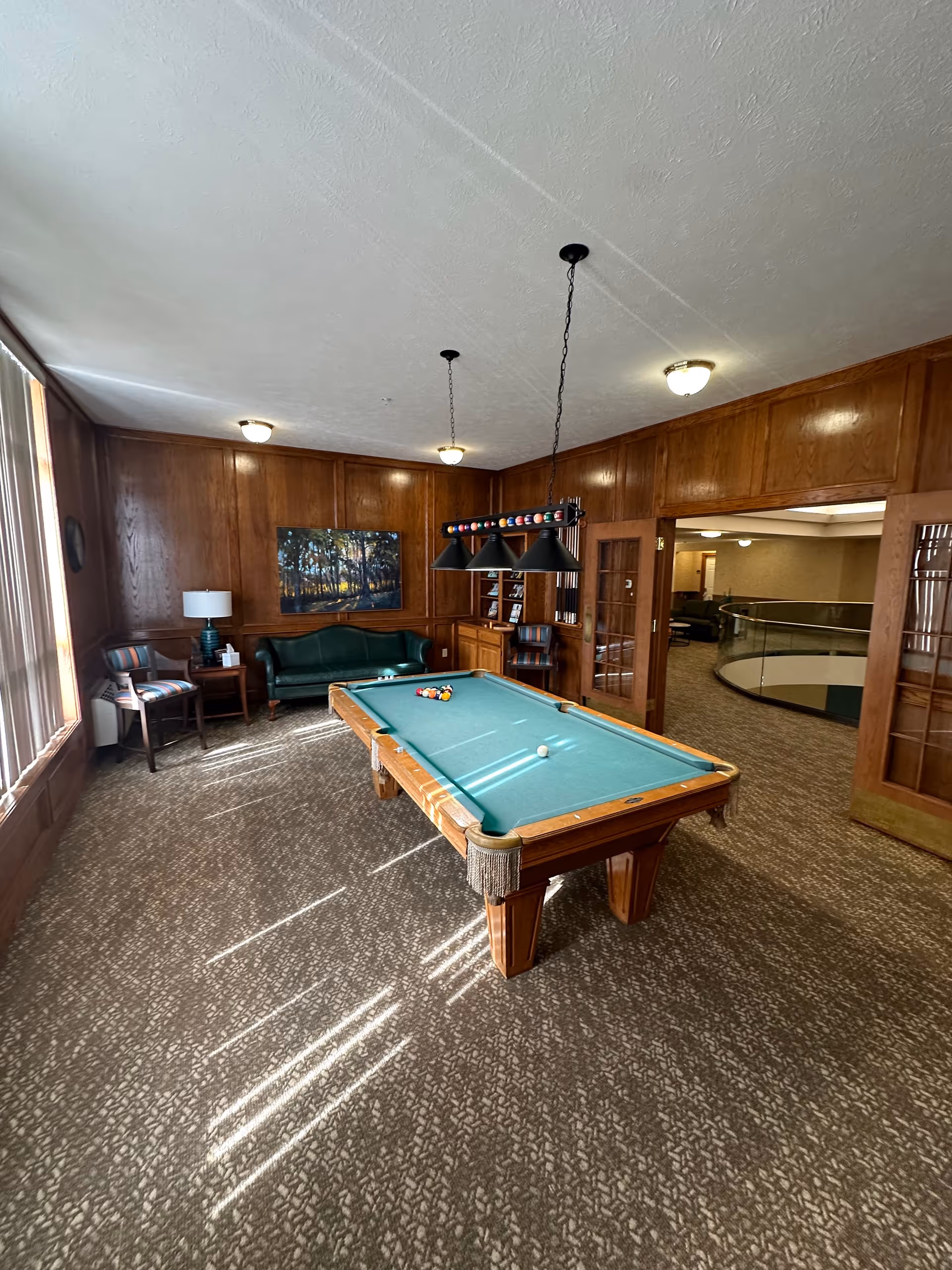 A well-lit room with wood-paneled walls featuring a pool table in the center with billiard balls arranged on it. There is a green leather couch and two striped chairs near a window with sunlight streaming in. A painting of a forest scene hangs on the wall above the couch. The room has carpeted flooring and a hanging light fixture above the pool table. Double glass doors open to an adjacent area with a curved railing.
