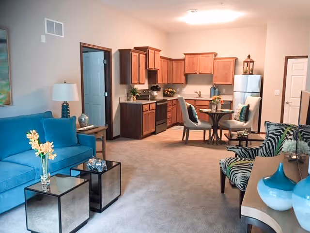 A cozy open-concept living area in a retirement community apartment featuring a blue sofa with a flower vase on a side table, two mirrored coffee tables, a patterned armchair, and a small dining table with two chairs. The kitchen area has wooden cabinets, a stove, a refrigerator, and a countertop with decorative items.