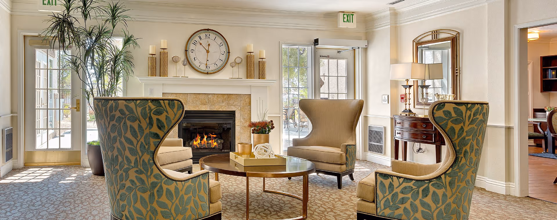 A cozy living room area in Heritage Springs featuring a fireplace with a clock and decorative candles on the mantel. The room has two patterned armchairs and one plain armchair arranged around a round wooden coffee table. There are large windows and glass doors letting in natural light, with plants and a side table with a lamp and mirror in the background.
