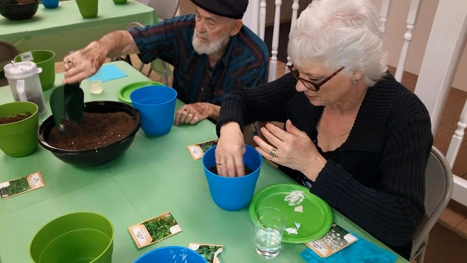 Two elderly individuals sitting at a table covered with a green tablecloth, engaging in a planting activity. One person is scooping soil into a blue pot, while the other is placing something into another blue pot. Several green and blue pots, seed packets, and a glass of water are on the table.