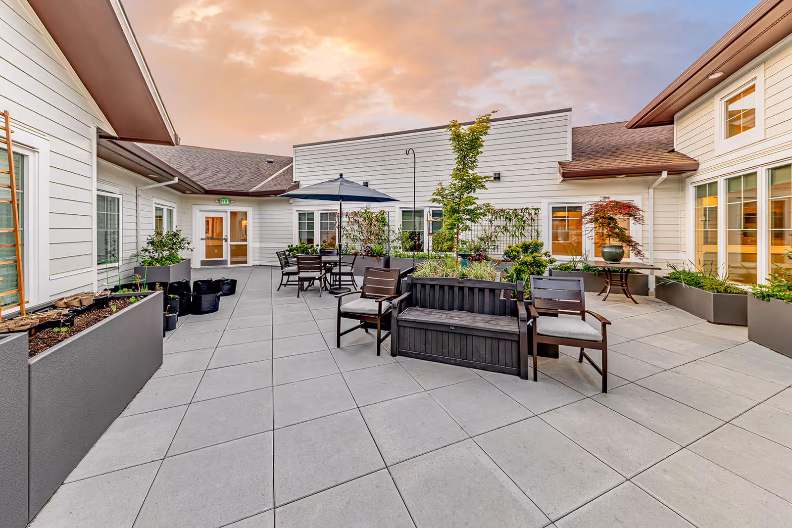 Outdoor patio area at Courtyard At Mount Tabor with tiled flooring, several planters with greenery, a black bench with cushions, two chairs, a table with an umbrella, and surrounding white buildings under a partly cloudy sky at sunset.