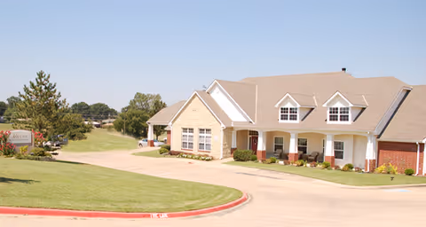 Exterior view of a single-story senior living facility building with a beige roof, brick and stone facade, and a covered porch with seating. The building is surrounded by a well-maintained lawn and a curved driveway. Trees and a clear blue sky are visible in the background.