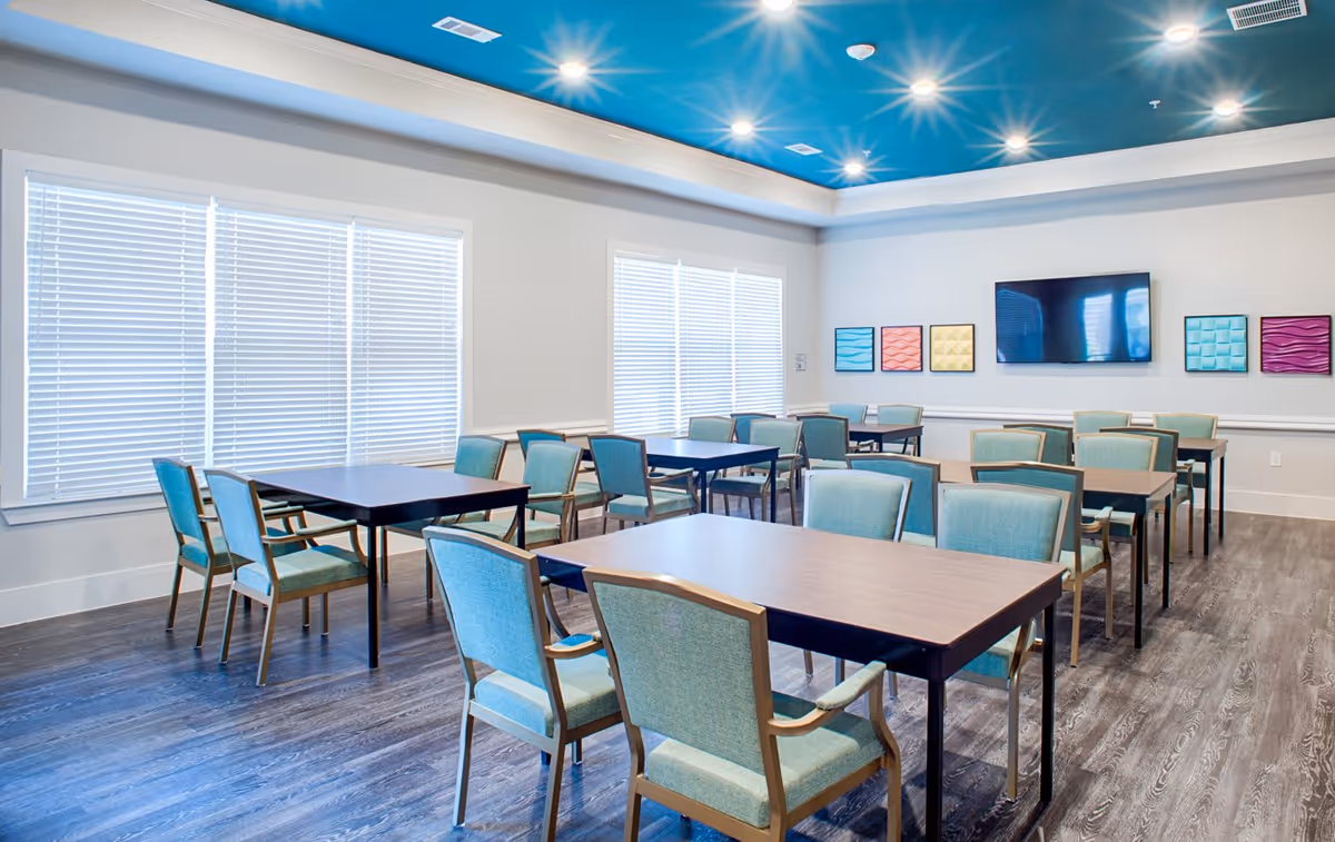 A bright and modern dining room with multiple tables and teal upholstered chairs arranged neatly. The room features large windows with white blinds, a blue ceiling with recessed lighting, and colorful square wall art. A flat-screen TV is mounted on the wall.