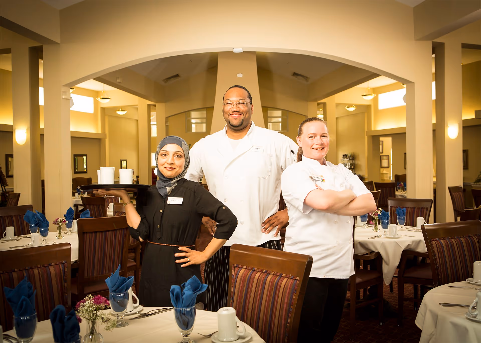 Three smiling staff members standing in a dining room of an assisted living facility. One woman wearing a hijab holds a tray with cups, a man in a chef's coat stands in the middle, and another woman in a chef's coat stands with arms crossed. The dining room has tables set with white tablecloths, blue napkins, glasses, and small flower arrangements.