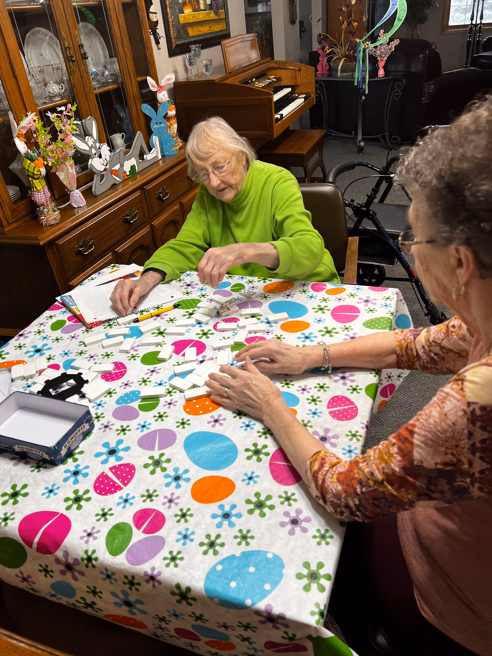 Two older women sit at a brightly patterned table in a communal room playing dominoes, with a piano and china cabinet in the background.