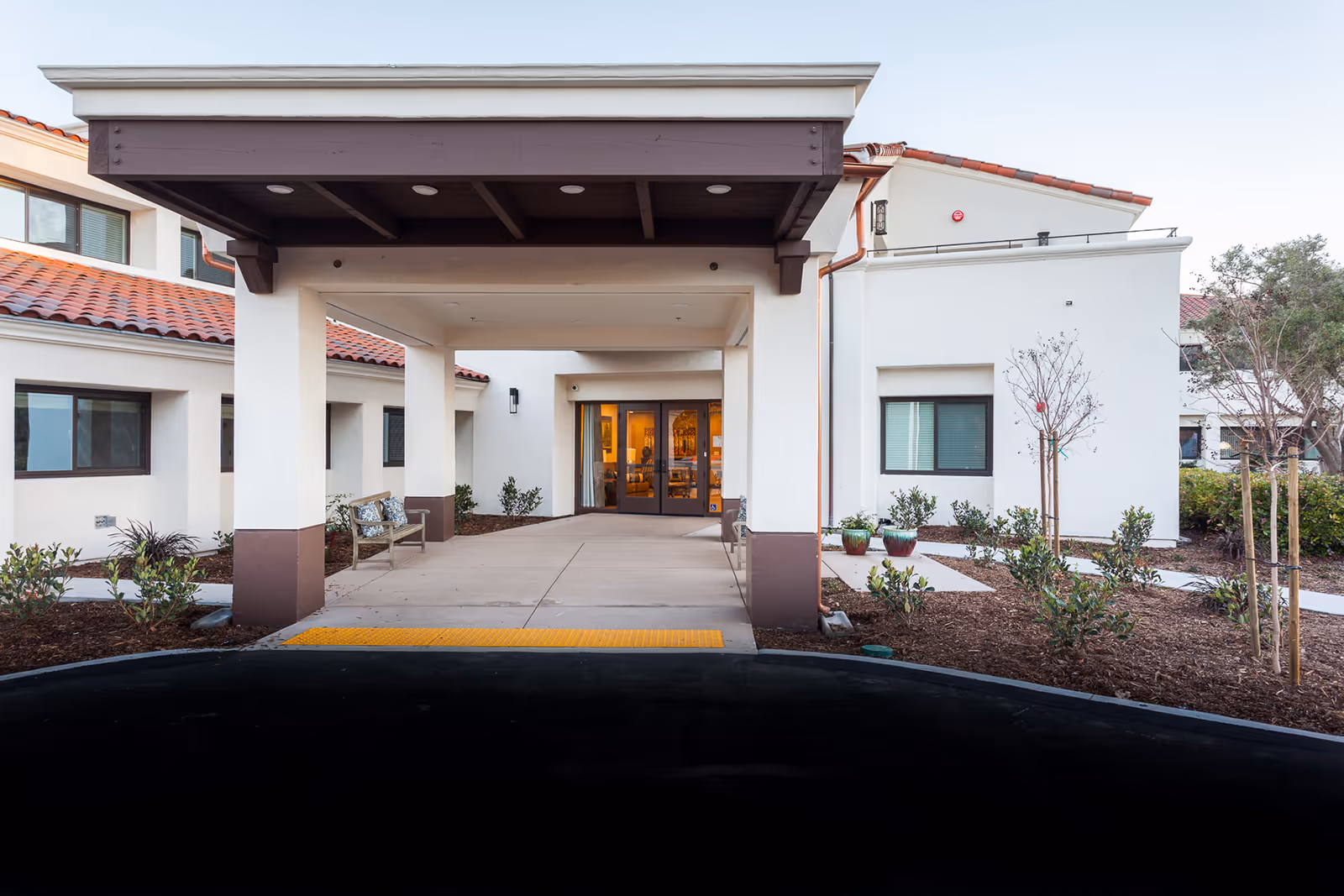 Covered drive-up entrance and canopy of a white stucco senior living facility with glass double doors and landscaped planting beds.