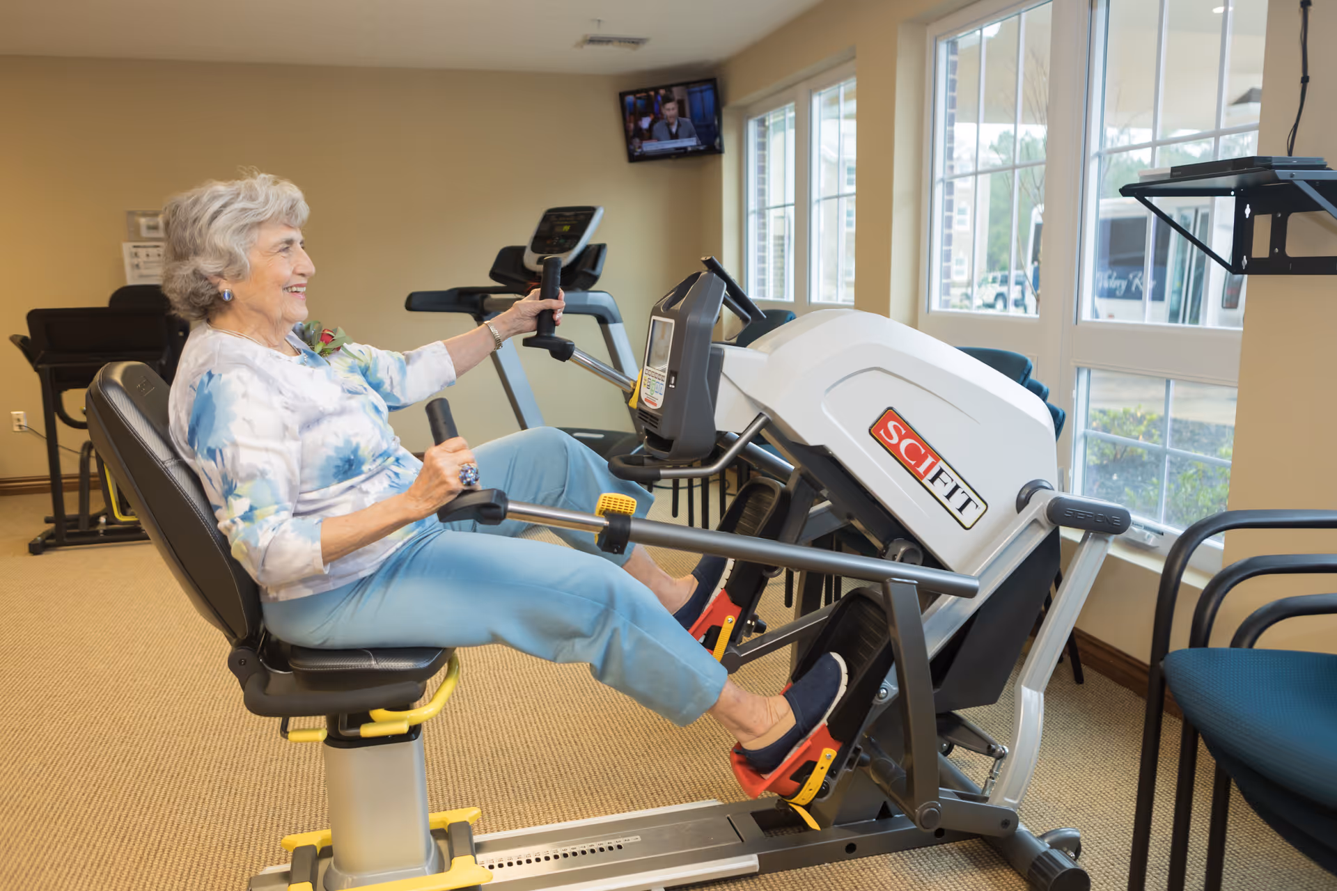 An elderly woman with gray hair is exercising on a recumbent bike in a well-lit fitness room with large windows. She is smiling and wearing a floral top and light blue pants. There is other exercise equipment visible in the background and a small TV mounted on the wall.