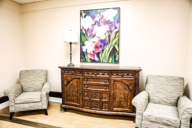Two upholstered armchairs flank an ornate wooden sideboard topped with a lamp and a colorful floral painting on a neutral wall.