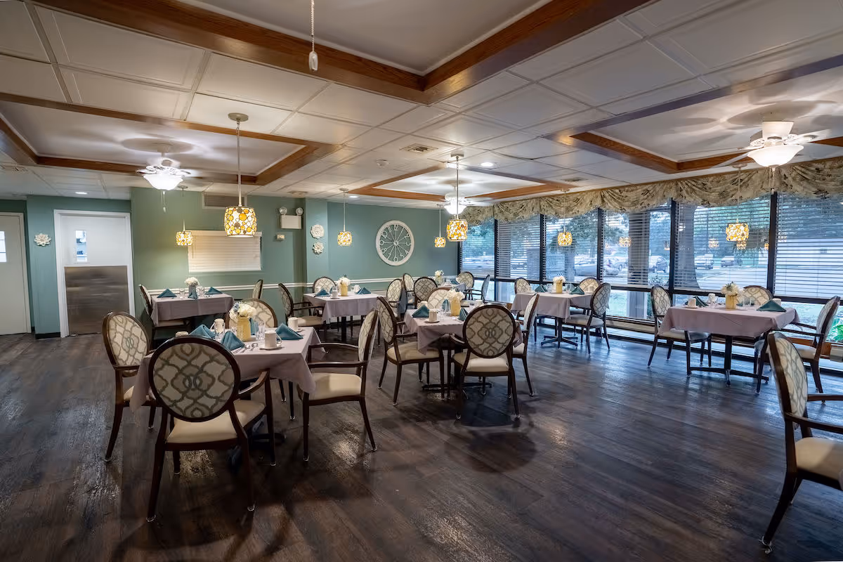 A dining room with multiple tables covered in light purple tablecloths, each set with teal napkins, white cups, and small flower arrangements. The room has large windows with blinds and floral valances, pendant lights hanging from the ceiling, and ceiling fans. The floor is dark wood, and the walls are painted a soft green with decorative wall accents and a large clock.
