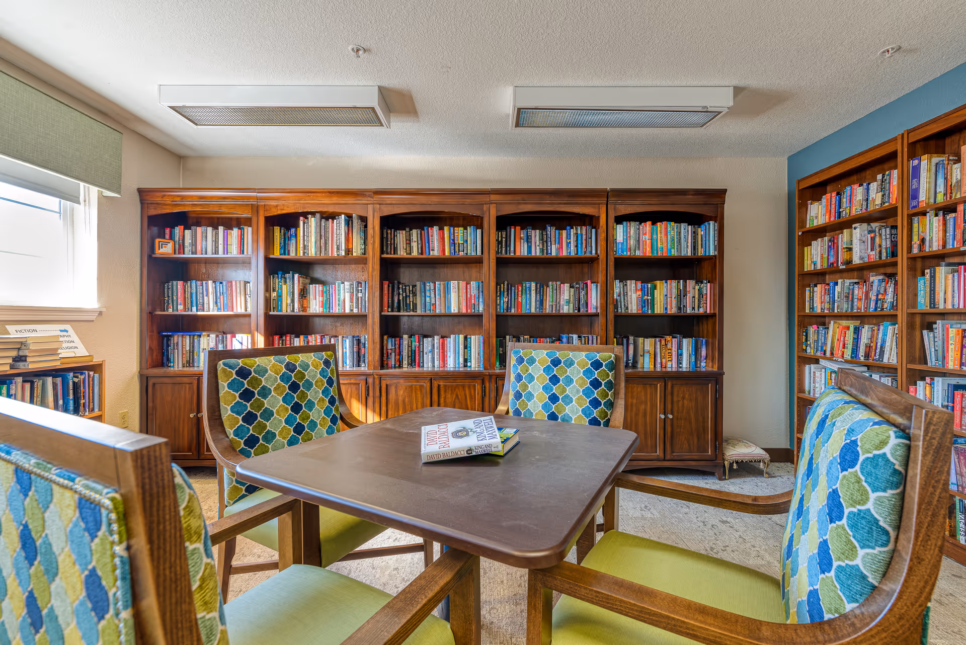 A cozy library room with wooden bookshelves filled with books along the walls. In the center, there is a square wooden table with four cushioned chairs featuring a blue, green, and beige patterned upholstery. Two books are placed on the table. A window with a green valance allows natural light into the room.