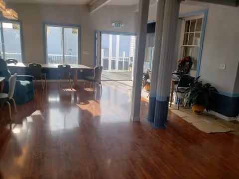 Spacious sunlit common room with wooden floors, tables and chairs near windows and an open doorway to a porch.