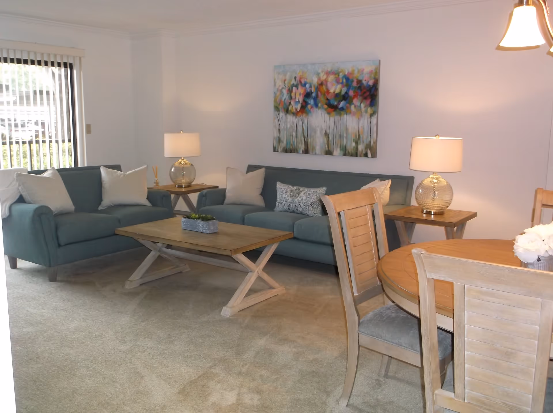 A living room with two teal sofas adorned with white and patterned pillows, a wooden coffee table in the center, two side tables each with a lamp, a colorful abstract painting on the wall, and a wooden dining table with chairs in the foreground.