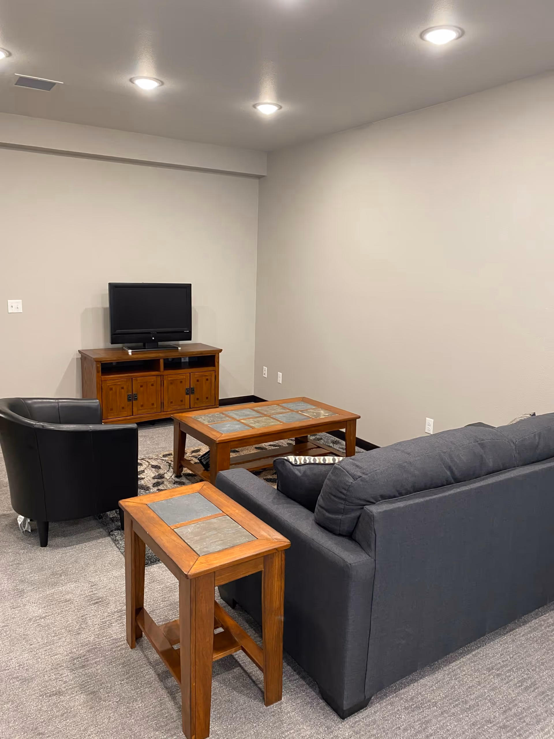 A small living room area with a dark gray couch, a black armchair, a wooden coffee table with tile inlays, a matching wooden side table, and a wooden TV stand with a flat-screen TV. The room has gray carpet, beige walls, and recessed ceiling lights.
