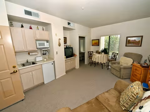 Interior view of a living area in an assisted living facility showing a small kitchenette with cabinets, microwave, sink, and mini fridge on the left. In the middle and right side, there is a small round dining table with two chairs near a window, a recliner chair, a wooden side table with a lamp, and a couch with a decorative pillow in the foreground. The walls are light-colored and there are framed pictures hanging on the walls.