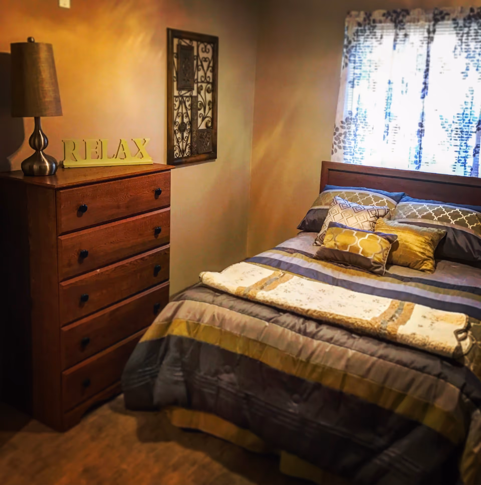 A cozy bedroom with a wooden bed covered in a striped comforter and multiple decorative pillows. Next to the bed is a wooden chest of drawers with a lamp and a decorative sign that says 'RELAX'. A window with patterned curtains allows natural light into the room, and a decorative wall piece hangs above the chest of drawers.