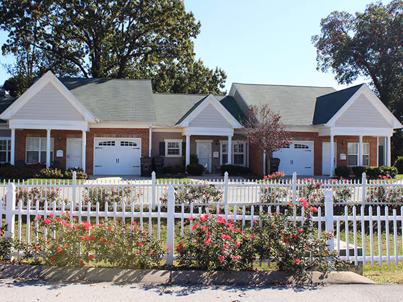 Exterior view of a single-story senior living facility building with brick walls, white trim, and green shingled roofs. The building has two garage doors and several windows. In front, there is a white picket fence with blooming red and pink flowers and some trees in the background under a clear blue sky.