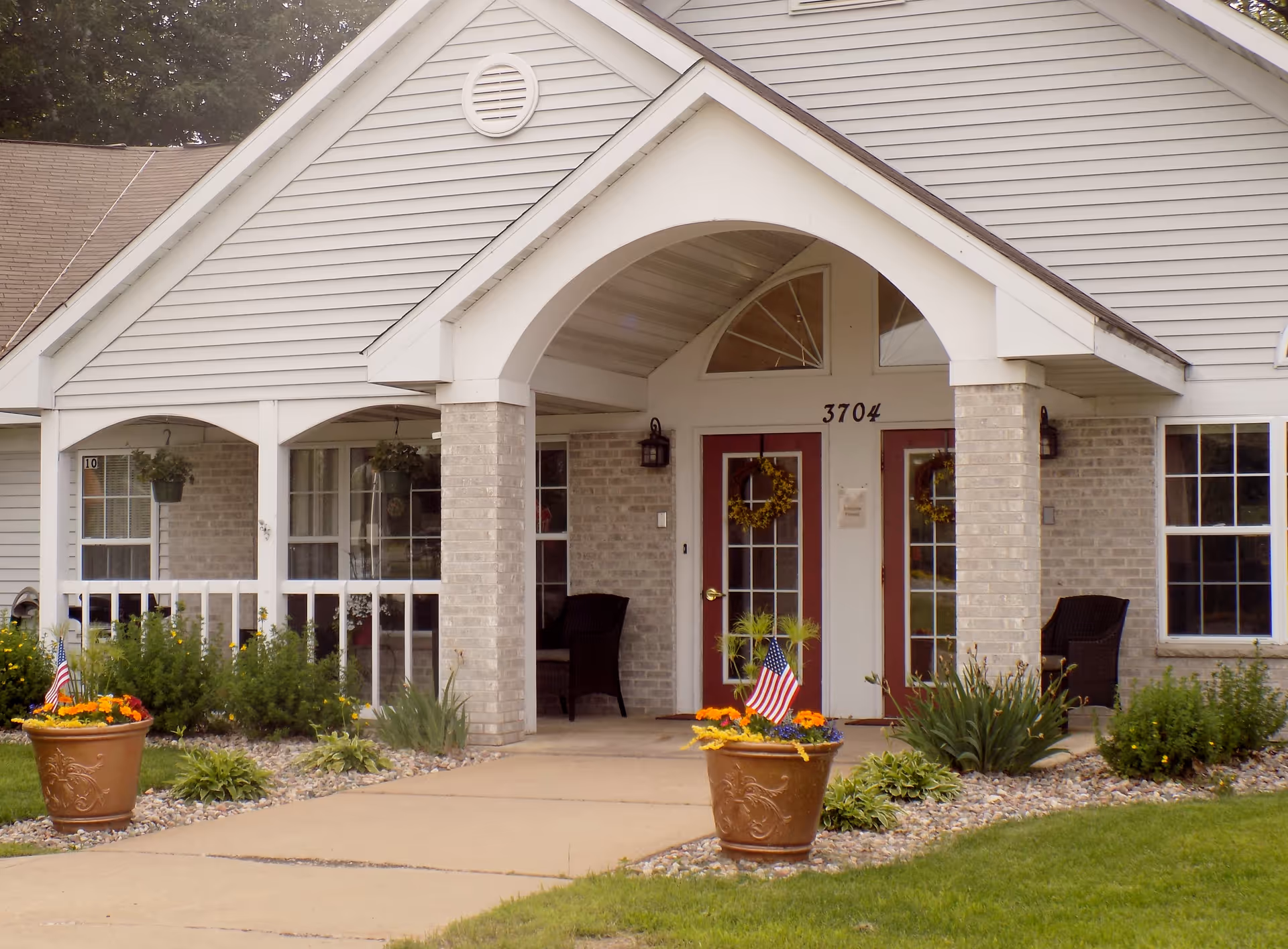 Front entrance of a senior living building with a covered porch, columns, double red doors numbered 3704, and potted flowers with American flags.