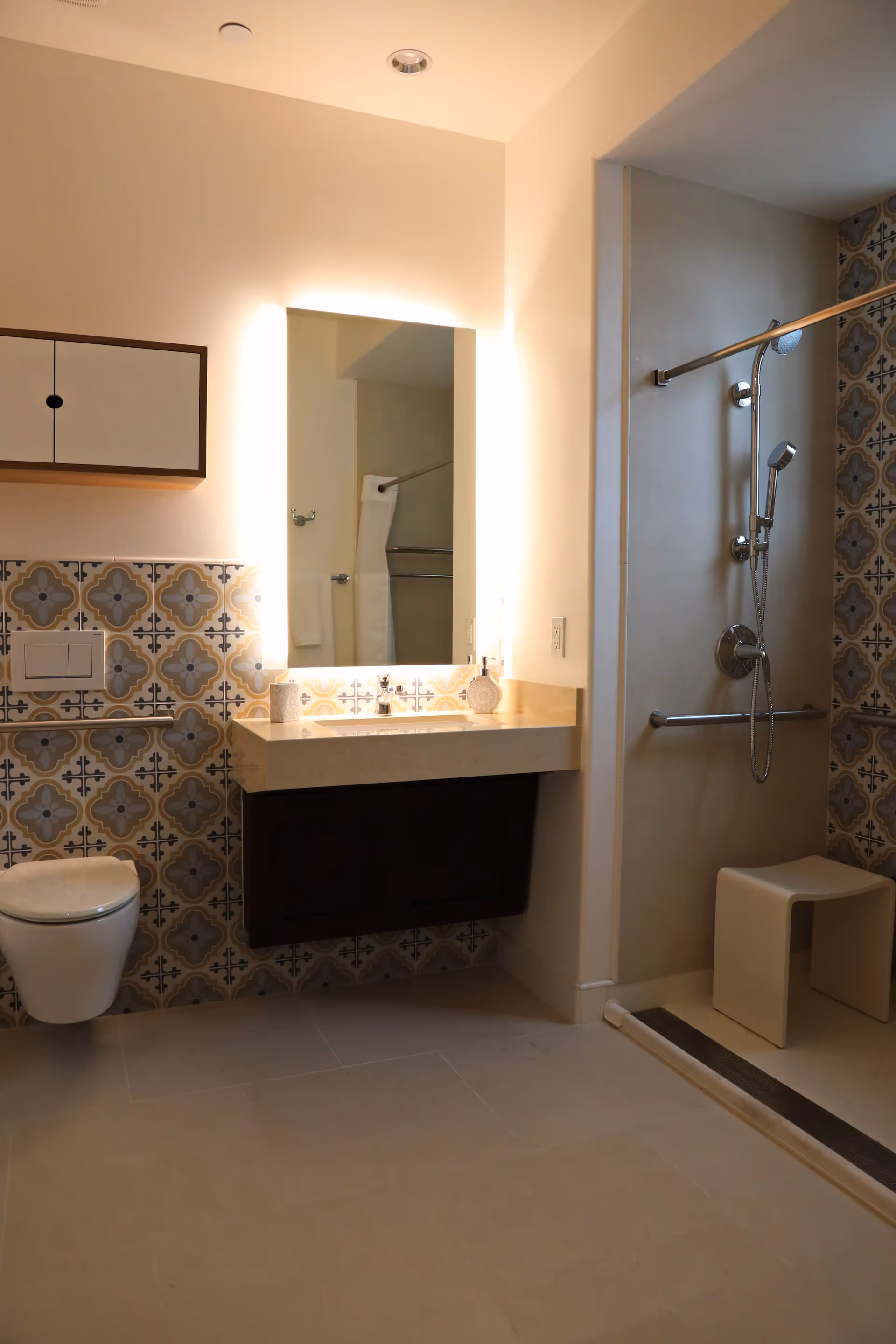 A modern bathroom featuring a wall-mounted toilet, a floating vanity with a sink, and a large illuminated mirror above it. The walls have decorative patterned tiles halfway up, and there is a walk-in shower area with a handheld showerhead, grab bars, and a white shower stool.